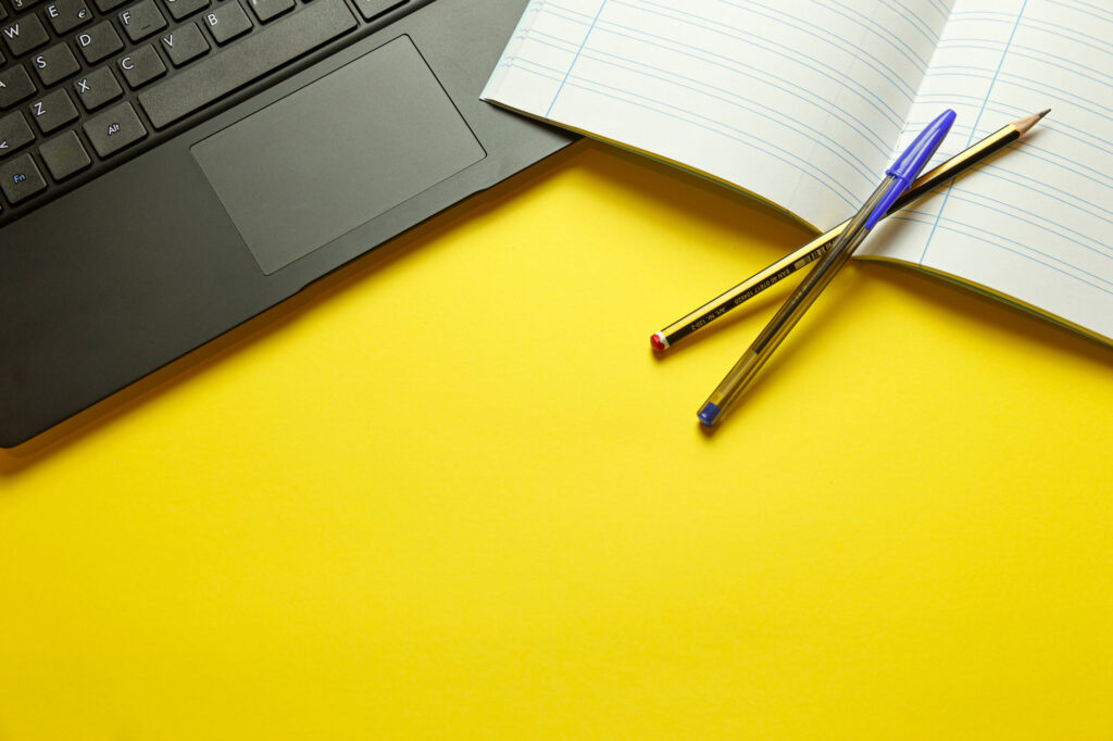 An open laptop and notebook with pen and pencil on top of a yellow table.
